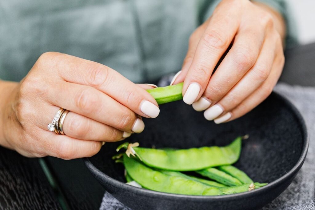 A woman with manicured nails shells fresh snap peas into a black bowl.